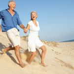 Senior Couple Enjoying Beach holding hands, running Down sand dune.