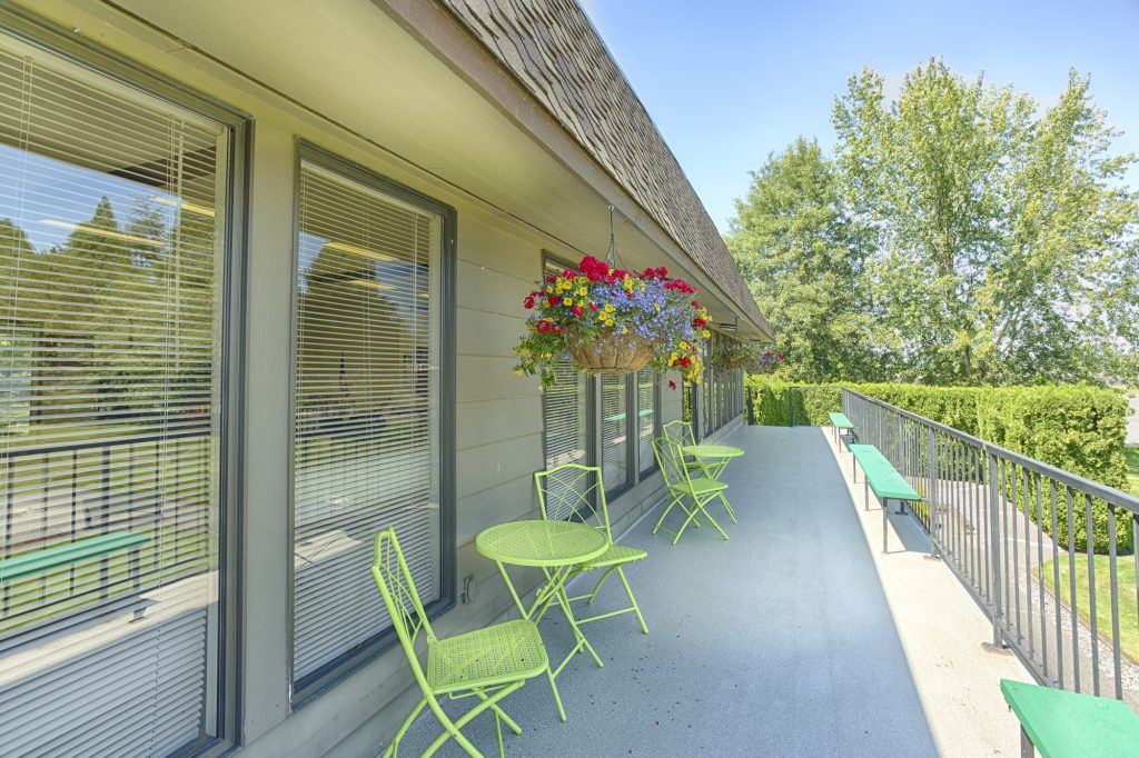 Long balcony outside the community center that overlooks the open outdoor area. Colorful flower bouquets hang from awning with bright green 3-piece seating sets are arranged below along the balcony.