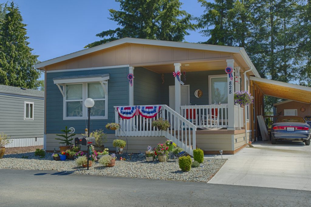 Beautiful manufactured home with small staircase leading up to covered porch. Painted with a shade of blue and decorated with an American flag theme. Side carport for one car coverage.