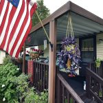 A resident proudly displays the American flag from their front porch/ Lush landscaping with 2 hanging flower pots. One with beautiful purple flowers. Small shed in the back.