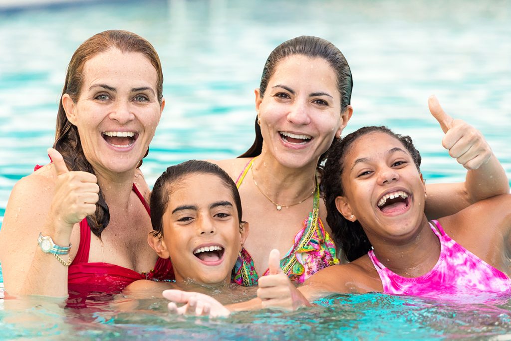 A couple of mothers posing with their son and daughter playing in a swimming pool. Giving thumbs up.