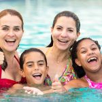 A couple of mothers posing with their son and daughter playing in a swimming pool. Giving thumbs up.