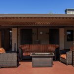 Comfortable outdoor seating with cushions situated under shaded area outside the clubhouse. One large couch and two single seats surround a small table.