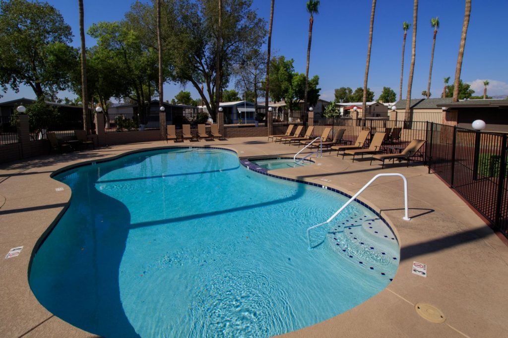 Large community pool with attached spa under the blue skies. Enclosed by black fencing and palm trees.