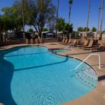 Large community pool with attached spa under the blue skies. Enclosed by black fencing and palm trees.